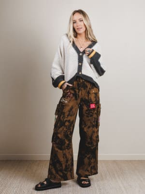 Woman wearing a white cardigan and camouflage pants with a red tag indoors.