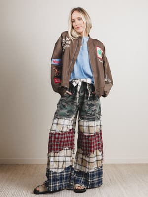 Person wearing a brown jacket with patches and plaid patchwork pants against a plain background