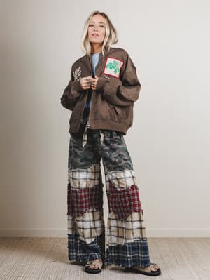 Woman wearing a brown jacket with patches and plaid pants against a plain background