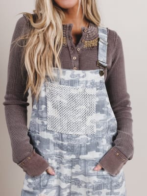 Person wearing a patterned denim overalls with a brown long-sleeve shirt underneath on a plain background
