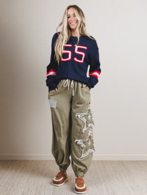 Woman wearing a navy sweater with red accents and green pants with floral patches, standing against a plain background.