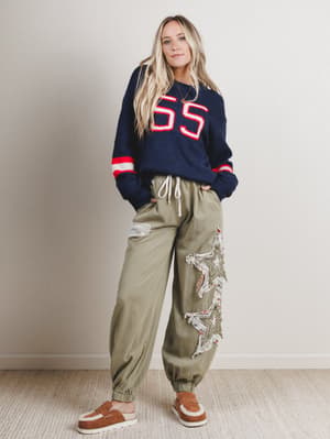 Woman wearing a navy sweater with red and white details and green pants with embroidery, standing against a plain background.