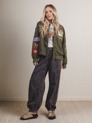 Woman wearing a green bomber jacket with patches and patterned pants against a plain background