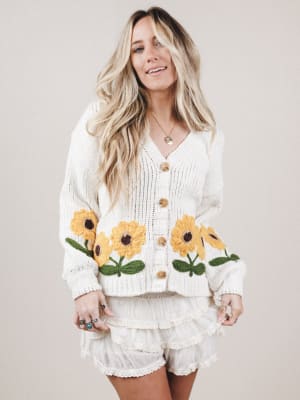 Woman wearing a white cardigan with sunflower patterns on a plain background
