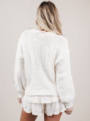 Woman wearing a white ribbed sweater and ruffled shorts on a plain background