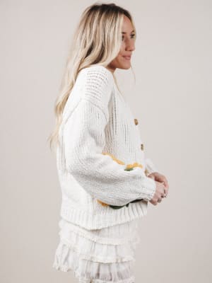 Woman wearing a white knitted cardigan and skirt with yellow accents on a plain background