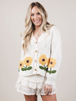 Woman wearing a white cardigan with sunflower designs on a plain background