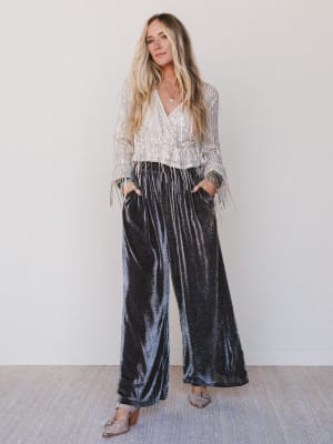 Woman wearing a patterned blouse and black velvet pants against a white wall.
