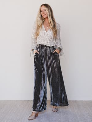 Woman wearing a patterned blouse and black velvet pants against a white wall.