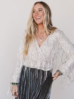 Woman wearing a sparkling silver top with fringe details against a plain background