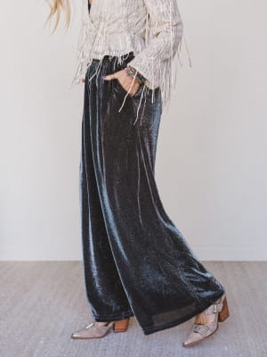 Person wearing black velvet wide-leg pants with a white top and brown shoes on a neutral background