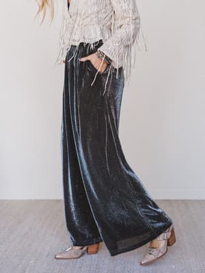 Person wearing black velvet wide-leg pants with a white top and brown shoes on a neutral background
