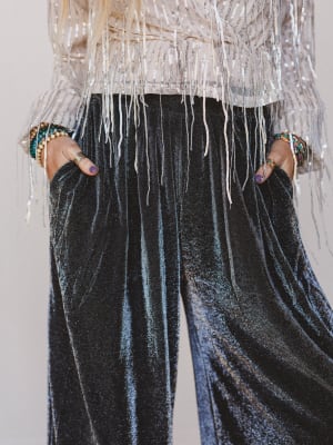 Person wearing black velvet pants with a white top with fringe details on a plain background