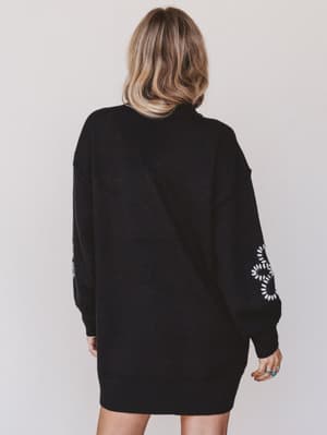 Person wearing a black sweater dress with white patterns on sleeves against a plain background