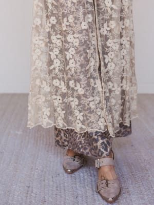Person wearing a long floral lace dress with leopard print pants and beige sandals on a textured floor.