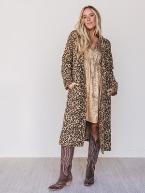 Woman wearing a leopard print coat over a gold dress with brown cowboy boots on a white background