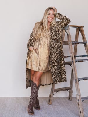 Woman in leopard print coat and dress with cowboy boots standing next to a ladder.