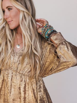 Woman wearing a gold dress with long blonde hair and multiple bracelets on a plain background