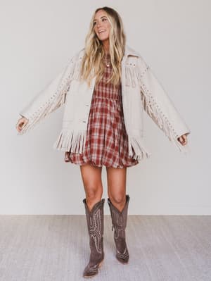 Woman wearing a plaid dress with a white fringe jacket and cowboy boots on a plain background
