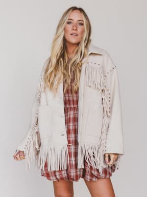 Woman wearing a beige fringed jacket over a plaid dress on a plain background