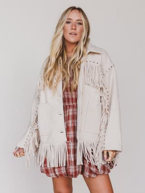 Woman wearing a beige fringed jacket over a plaid dress on a plain background