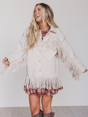 Woman wearing a beige fringed jacket over a plaid dress on a plain background