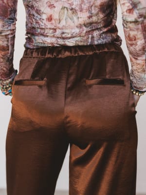 Brown satin pants worn by a person with a patterned top and colorful bracelets on a light background