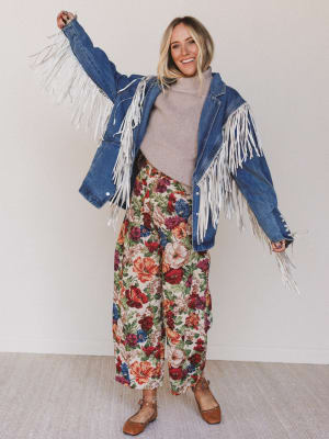 Woman wearing a denim jacket with white fringe, gray turtleneck, and floral pants against a plain background