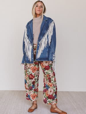Woman wearing a blue denim jacket with white fringe, beige sweater, and floral pants on a plain background