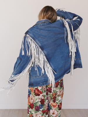 Person wearing a blue denim jacket with white fringe, standing against a plain background
