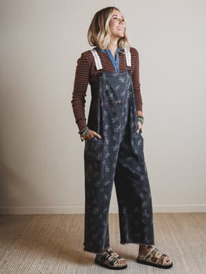 Person wearing floral overalls and a striped shirt against a plain background
