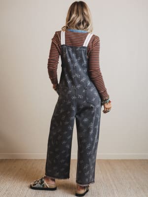 Person wearing floral overalls and a striped shirt against a plain wall.