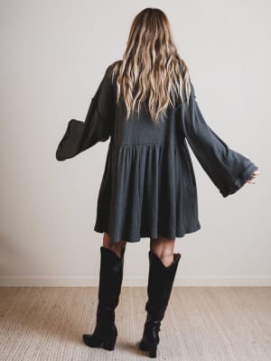 Person wearing a black dress with long sleeves and black knee-high boots in a minimal indoor setting.