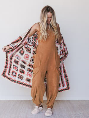 Woman wearing a brown jumpsuit with a patterned shawl draped over her shoulders against a white background.
