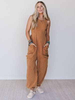 Woman wearing a rust sleeveless jumpsuit, colorful bracelets, and white shoes against a white background