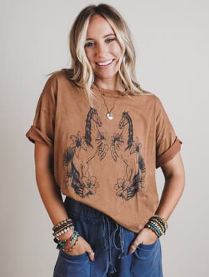 Woman wearing a brown t-shirt with horse designs and blue jeans, smiling against a plain background.