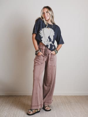 Woman wearing a graphic t-shirt and brown pants standing against a plain wall.