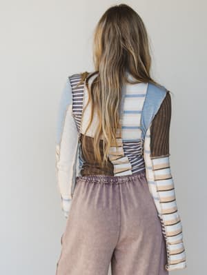 Back view of a woman wearing a colorful striped sweater and brown pants against a plain background