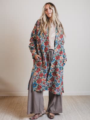 Woman wearing a long floral coat over a white shirt and gray pants on a plain background