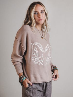 Woman wearing a beige sweater with a horse design on a plain background