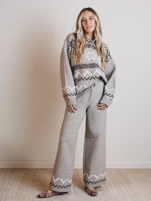 Woman wearing a gray patterned sweater and pants standing against a plain wall.