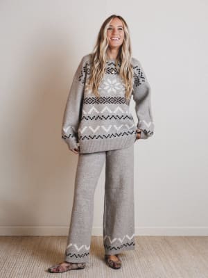 Woman wearing a gray patterned sweater and pants standing against a plain wall.