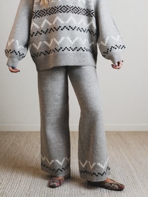 Person wearing a gray sweater and pants with a pattern, standing on a carpeted floor.