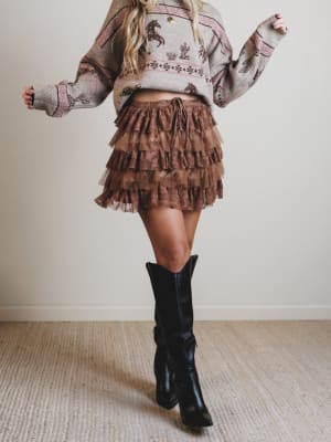 Person wearing a patterned sweater and tiered skirt with black knee-high boots on a plain background