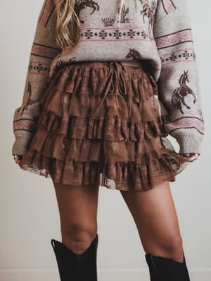 Person wearing a patterned sweater and brown tiered skirt with black boots on a plain background