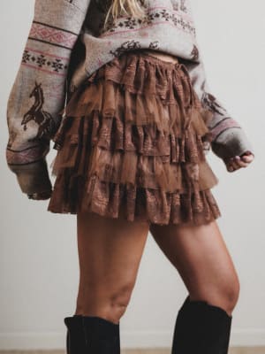 Person wearing a patterned sweater and brown ruffled skirt with black knee-high boots.