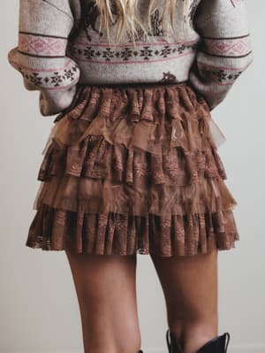 Person wearing a patterned sweater and brown ruffled skirt on a plain background