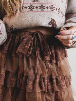 Person wearing a patterned sweater and brown ruffled skirt with a blurred background