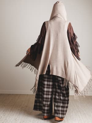 Person wearing a beige hooded vest with brown accents and plaid pants in a neutral setting