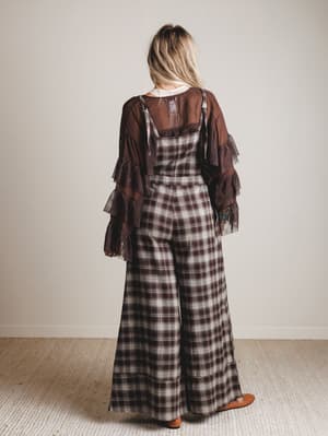 Person wearing a plaid jumpsuit with a brown shawl over shoulders against a plain background